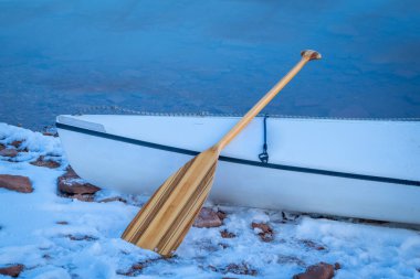 bow of a decked expedition canoe with a wooden paddle on a lake shore in winter clipart