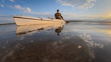 Gün batımında sakin bir gölde yaşlı bir kano kürekçisinin alçak açılı görüntüsü.