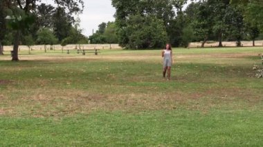 Beyaz kız parkta dikiliyor ve gri elbiseli kameraya doğru koşuyor.