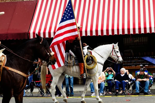 観客と小さな町のパレードで白い馬に乗っているアメリカの旗を持っている男 — ストック写真