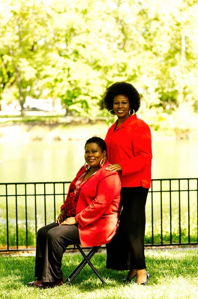 Two Middle Aged African American Women Outdoors In Black Pants And Red Jackets One Sitting And One Standing