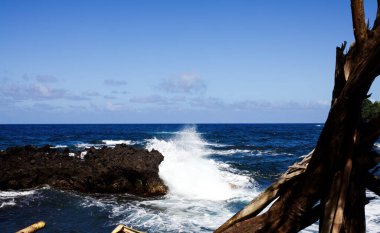 Büyük Hawaii adası mavi okyanusu ve gökyüzünü beyaz bulutlar ve kara lav kayasına çarpan dalgalarla seyrediyor.