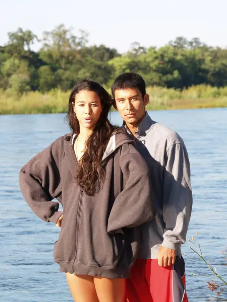 Man And Woman Couple Standing At River Looking At Camera With Green Plants And Trees In Background