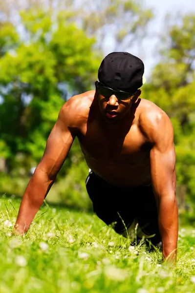 非洲裔美国人没有衬衫帽和短裤在公园绿草和浓重的阳光下做俯卧撑 图库图片