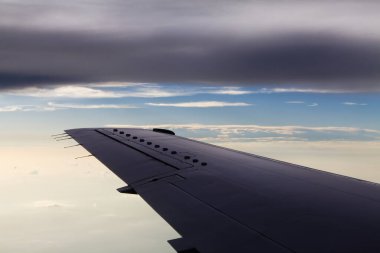 Ticari uçak kanatları, üzerinde kara bulutlar ve altında mavi gökyüzü olan beyaz katmanlarla uçuyor.