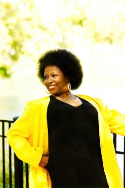 Çekici gülümseyen orta yaşlı Afro-Amerikan kadın açık havada sarı ceketli ve siyah elbiseli.