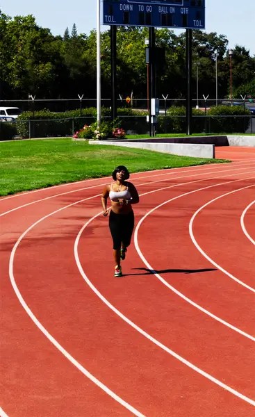 Pistte koşan genç Afrikalı Amerikalı kadın gri spor sutyen ve siyah pantolon giyiyor.