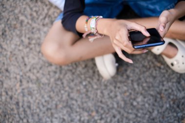Şort giyen Asyalı genç kız yerde bağdaş kurup akıllı telefonunu kullanmaktan zevk alıyor.