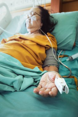 Sick Southeast Asian elderly female patient sleeping on hospital bed with oxygen and medical support. Open hand, oximeter placed on her thumb. clipart