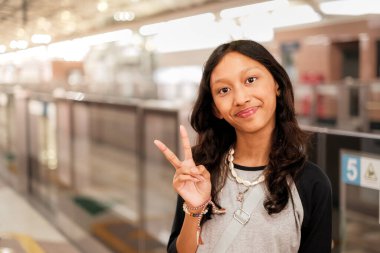 Güneydoğu Asyalı genç bir kız metro istasyonuna banliyö treninin varış zamanını beklerken mutlu ve neşeli görünüyor. Kameraya bakıp gülümsüyorsun..