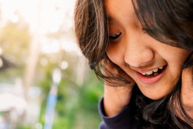 Mutlu şirin Asyalı genç kız, yakın çekimde aşağıya bakıyor ve uzun siyah saçlarına dokunarak samimi bir gülümseme takınıyor.