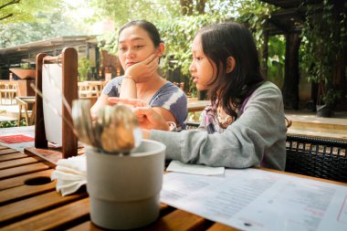 Asyalı anne ve kız restoran menüsüne bakıyor, öğle yemeği için yemekleri tartışıyor ve seçiyorlar.