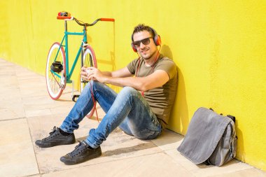 Rear image of a young man sitting on the floor leaning against a yellow wall listening to music through headphones while using a mobile phone. He has a fixie bike leaning against the wall.