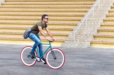 Side view of a young man is wearing sunglasses and carrying shoulder bag while riding fixie bike.