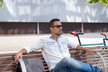 A young man sitting on a city bench relaxing, wearing sunglasses and looking into the future.