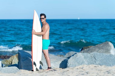 Hispanic surfer boy standing on the beach in neoprene holding his blue surfboard at sunset