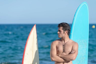 A Hispanic surfer guy standing on the beach in a swimsuit looking at the camera next to two surfboards.