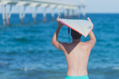 Back view of Hispanic surfer boy standing on the beach in swimsuit looking out to sea with surfboard on his head.