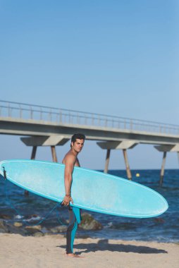 Hispanic surfer boy standing on the beach in neoprene holding his blue surfboard at sunset