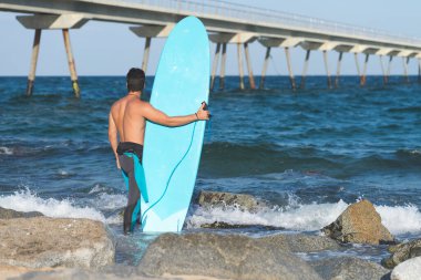 Hispanic surfer boy standing on the beach in neoprene holding his blue surfboard at sunset