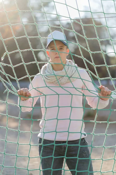 Teenager with cap behind a rope net in a park looking away.