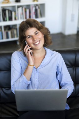 Telefonda konuşurken ve kameraya bakarken elinde dizüstü bilgisayarla koltukta oturan gülümseyen genç bir kadın..