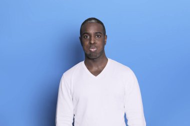 Mavi arka planda izole edilmiş beyaz süveter giyen neşeli, Afro-Amerikalı bir adamın portresi. Tasasız genç milenyum adamı kameraya bakıyor ve gülüyor.