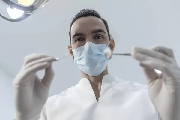 Low angle shot of a male dentist, doing the exam and checking up on oral health, teeth, and gums of the patient. The stomatologist holds dental instruments.
