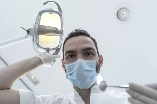 Low angle shot of a male dentist, doing the exam and checking up on oral health, teeth, and gums of the patient. The stomatologist holds dental instruments.