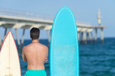 Neoprene mayo giymiş yakışıklı ve çekici bir sörfçü elinde sörf tahtası, yaz sporu ve tatillerin tadını çıkardıktan sonra havalı poz veriyor.