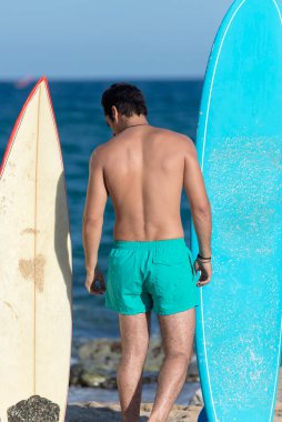 Neoprene mayo giymiş yakışıklı ve çekici bir sörfçü elinde sörf tahtası, yaz sporu ve tatillerin tadını çıkardıktan sonra havalı poz veriyor.