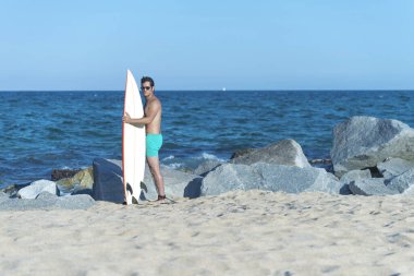 Neoprene mayo giymiş yakışıklı ve çekici bir sörfçü elinde sörf tahtası, yaz sporu ve tatillerin tadını çıkardıktan sonra havalı poz veriyor.