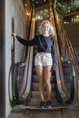 Gece mekanik merdivenlerde dururken elinde uzun bir tahtayla kameraya bakan gülümseyen bir kadının ön görüntüsü.