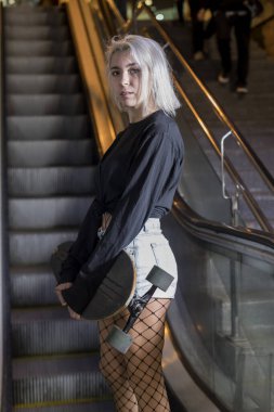 Gece mekanik merdivenlerde dikilirken elinde uzun bir tahta tutan şortlu bir kadının kamera görüntüsü.