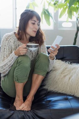 Rahat bir kadın telefonuna bakarken kahvesinden zevk alır, modern bir eğlence resmi. Sıcak bir örgü süvetere sarılmış ve rahat bir şekilde oturarak, güneşli bir odada huzurlu bir sabahın basitliğini temsil ediyor. Bu resim özünü yakalar. 