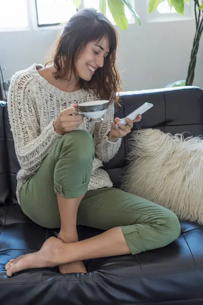 一个快乐的漂亮女人坐在沙发上 带着碗和智能手机 体现了舒适和现代生活 图库图片