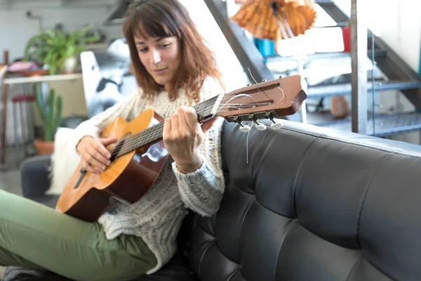 음악에 여성은 소파에 편안하게 앉아있는 기타를 연주합니다 그녀의 캐주얼 복장과 스톡 사진