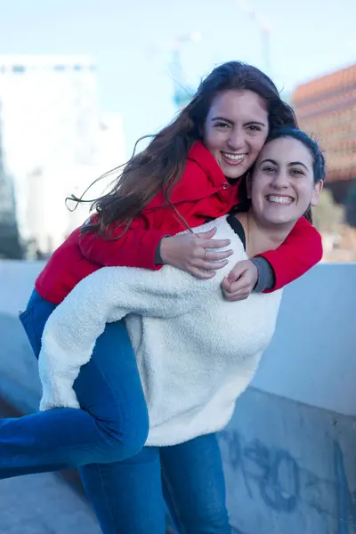 Two friends share a joyful piggyback moment, one in a red hoodie carrying the other in a white fluffy jacket, both smiling and having fun