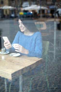 Mavi kazaklı genç bir kadın bir kafede telefonuna odaklanıyor. Önünde kahve fincanı ve çay tabağı var. Pencereden bakıyor.