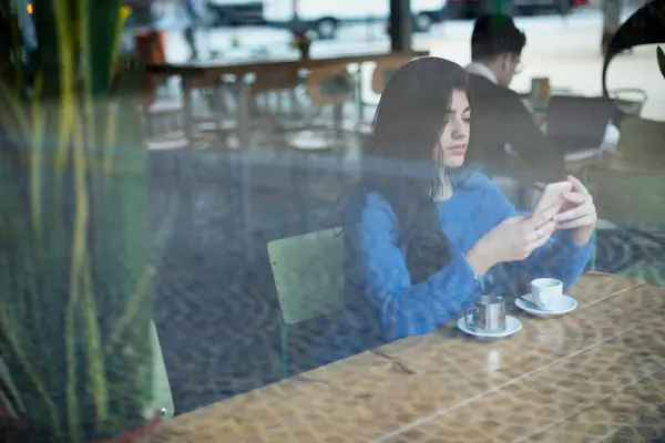 A young woman in a blue sweater focuses on her phone at a cafe, with a coffee cup and saucer in front of her, viewed through a window