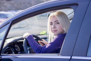 Mor bir süveter giyen genç sarışın bir sürücü, bir arabanın direksiyonunda güvenle oturuyor, rahat bir gülümsemeyle geriye bakıyor..