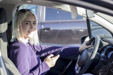 Mor kazaklı neşeli sarışın bir kadın arabanın içinde, bir elinde akıllı bir telefon, diğer elinde direksiyon simidi, kafasına güneş gözlüğü takıyor.