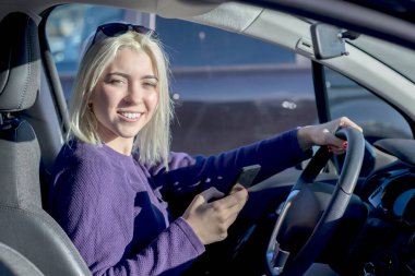 Mor kazaklı neşeli sarışın bir kadın arabanın içinde, bir elinde akıllı bir telefon, diğer elinde direksiyon simidi, kafasına güneş gözlüğü takıyor.