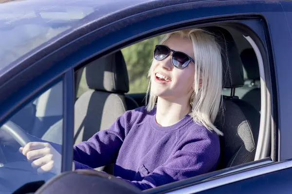 Güneş gözlüklü neşeli bir sarışın kadın, araba kullanıyor, gülüyor ve neşeli görünüyor, güneşli bir günde araba sürmenin zevkini gösteriyor.