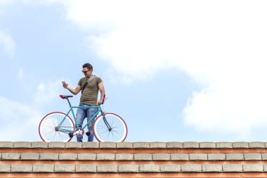 Çatıda renkli bir bisikletle genç bir adam akıllı telefonuna bakıyor, şehir yaşamını, bağlantısını ve aktif yaşam tarzını resmediyor.