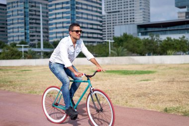 Şehir parkında fixie bisikleti süren modaya uygun genç bir adam. Arka planda modern binalar var. Çevre dostu bir seyahat örneği.