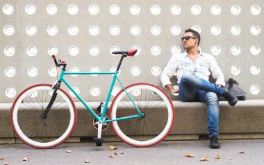 Gündelik giysiler içinde modaya uyan bir adam renkli sabit vitesli bisikletinin yanında dekoratif bir şehir duvarına karşı mola veriyor.