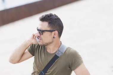 Güneş gözlüklü şık bir adam ve bir kurye çantası telefon görüşmesi yapıyor, şehir ortamında eski bir bisiklete yaslanıyor, hareketlilik ve şehir yaşamını öneriyor.