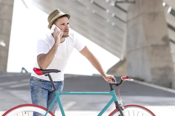 Young man in casual attire, talking on the phone while holding a stylish bicycle, set against an impressive urban structure, suggesting mobility and connectivity