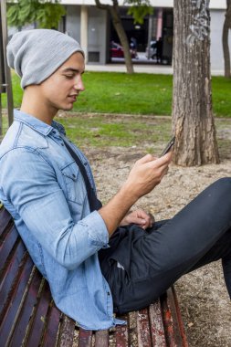 Kot ceket ve gri bere giymiş odaklanmış genç bir erkek, ağaçlar ve çimlerle çevrili akıllı telefonunu kullanarak bir bankta oturur..